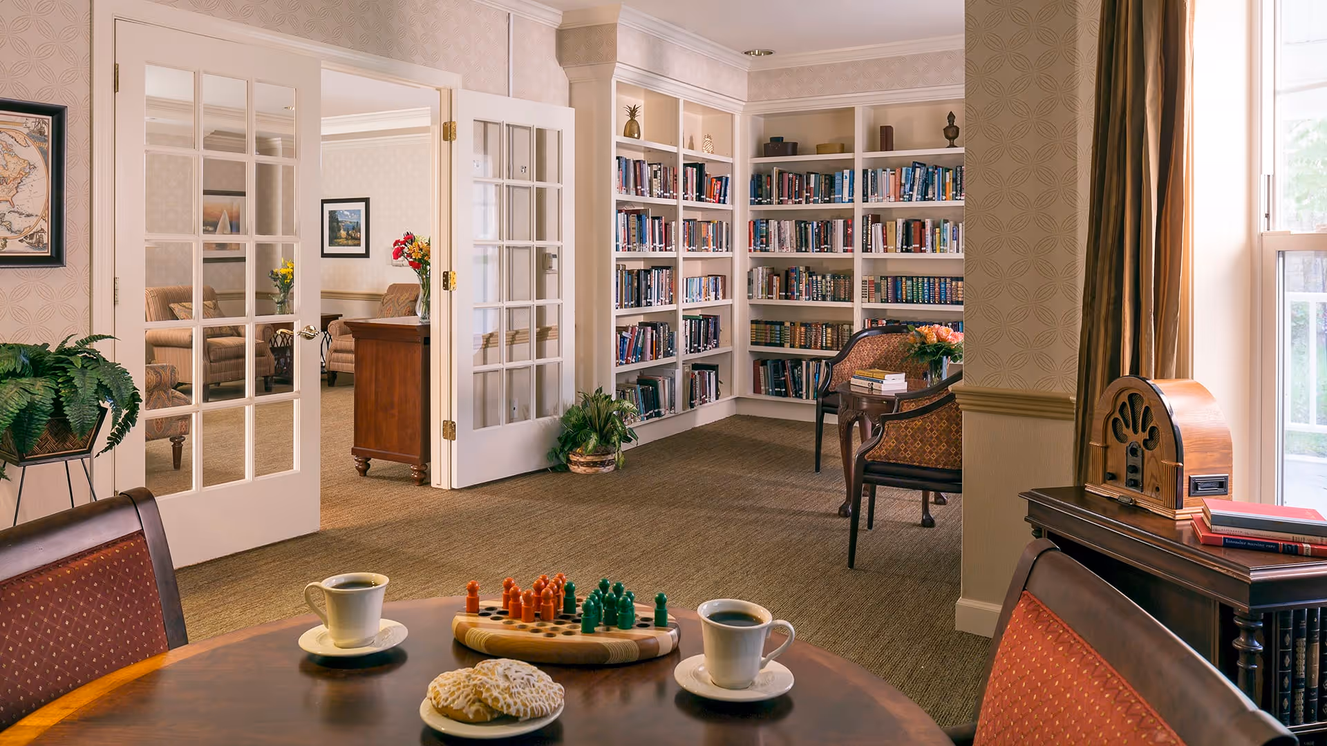 A cozy library and sitting area in a senior living facility with built-in bookshelves filled with books, a round table with two cups of coffee and a board game, comfortable chairs, and a vintage radio on a side table near a window with curtains. French doors open to an adjacent room with sofas and framed artwork on the walls.