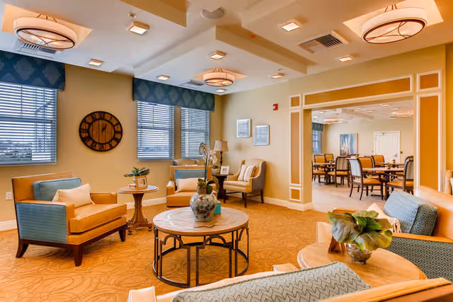 Bright, warmly decorated senior living lounge with upholstered chairs, round coffee tables, plants, and a view into an adjacent dining area.
