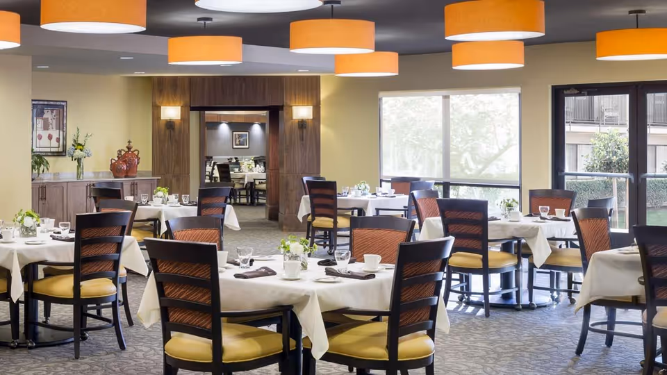 Bright dining room with round tables set with white tablecloths and wooden chairs under large orange pendant lights.