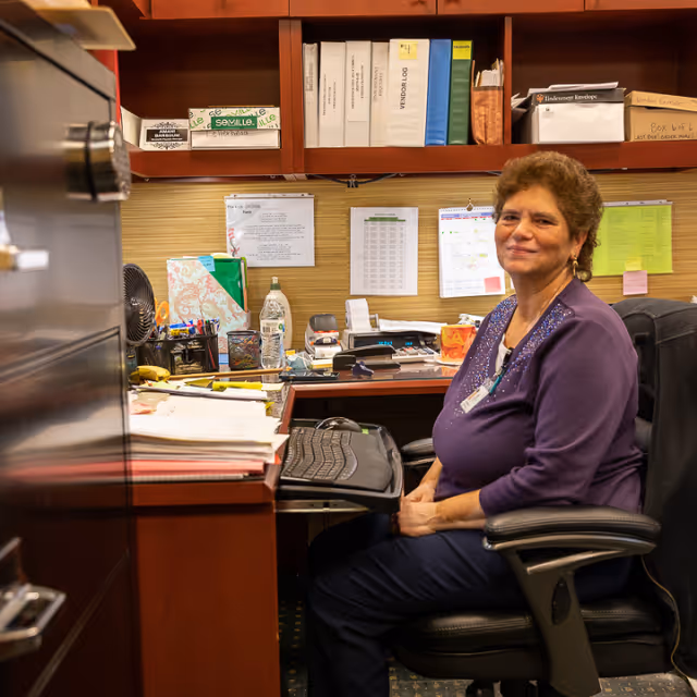 A woman with short curly hair wearing a purple top sits in an office chair at a desk with a keyboard, papers, and office supplies. Behind her are shelves with binders and boxes, and a bulletin board with pinned papers.