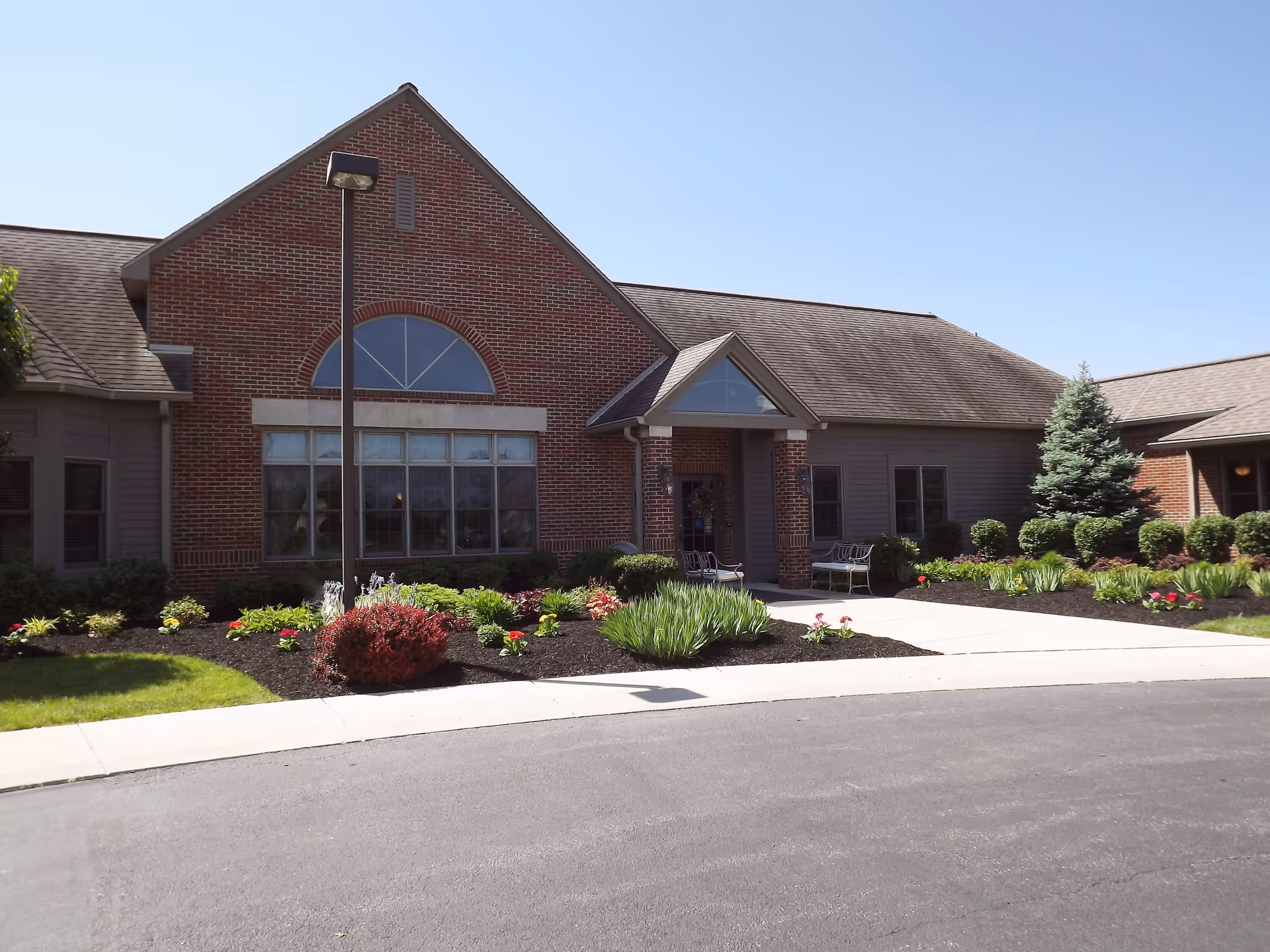 Exterior view of a single-story brick and siding building with a peaked roof, large windows, a covered entrance with benches, landscaped flower beds, shrubs, and a small tree under a clear blue sky.