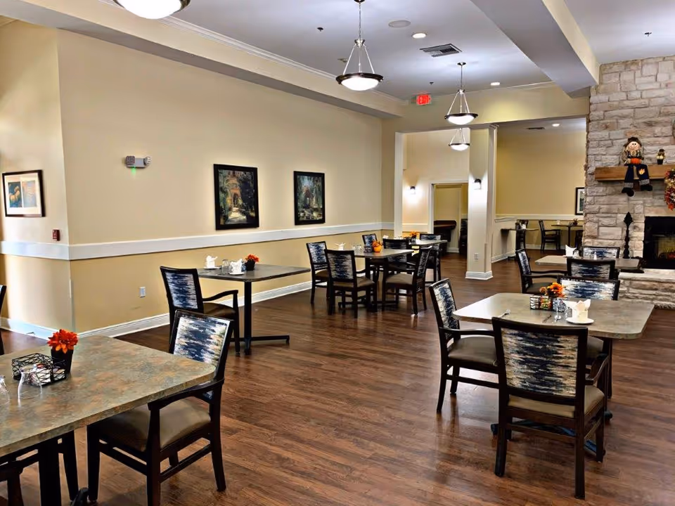 A dining area in an assisted living facility with several tables and chairs arranged on a wooden floor. The walls are painted beige and decorated with framed artwork. There is a stone fireplace on the right side with seasonal decorations. Ceiling lights hang above the tables, and small flower arrangements are placed on each table.