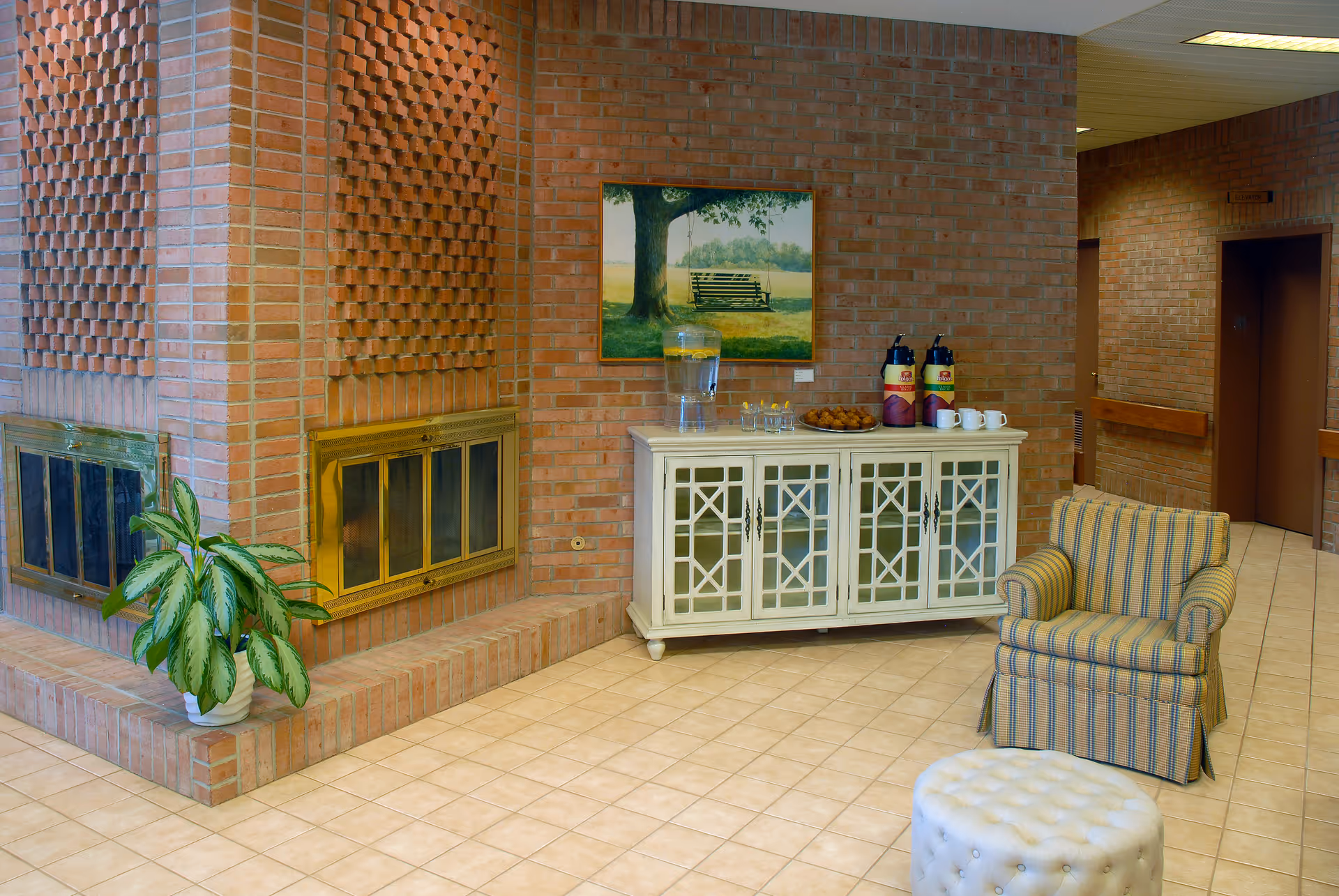 A cozy senior living facility common area with a brick fireplace featuring brass doors, a potted plant on the hearth, a white cabinet with glass doors holding a water dispenser, glasses, coffee dispensers, and mugs. A framed painting of a tree with a swing hangs on the brick wall above the cabinet. There is a striped armchair and a white tufted ottoman on a tiled floor. In the background, there is a hallway with an elevator door.