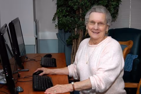 An elderly woman wearing glasses and a light pink sweater is sitting at a desk using a computer mouse and keyboard. There are two computer monitors on the desk, and a large green plant is visible in the background along with a dark green armchair.