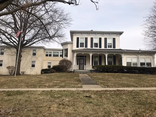 Front exterior view of a two-story building with white walls and black shutters, a covered porch with decorative columns, a flagpole with an American flag, leafless trees, and a grassy lawn with a concrete walkway leading to the entrance.