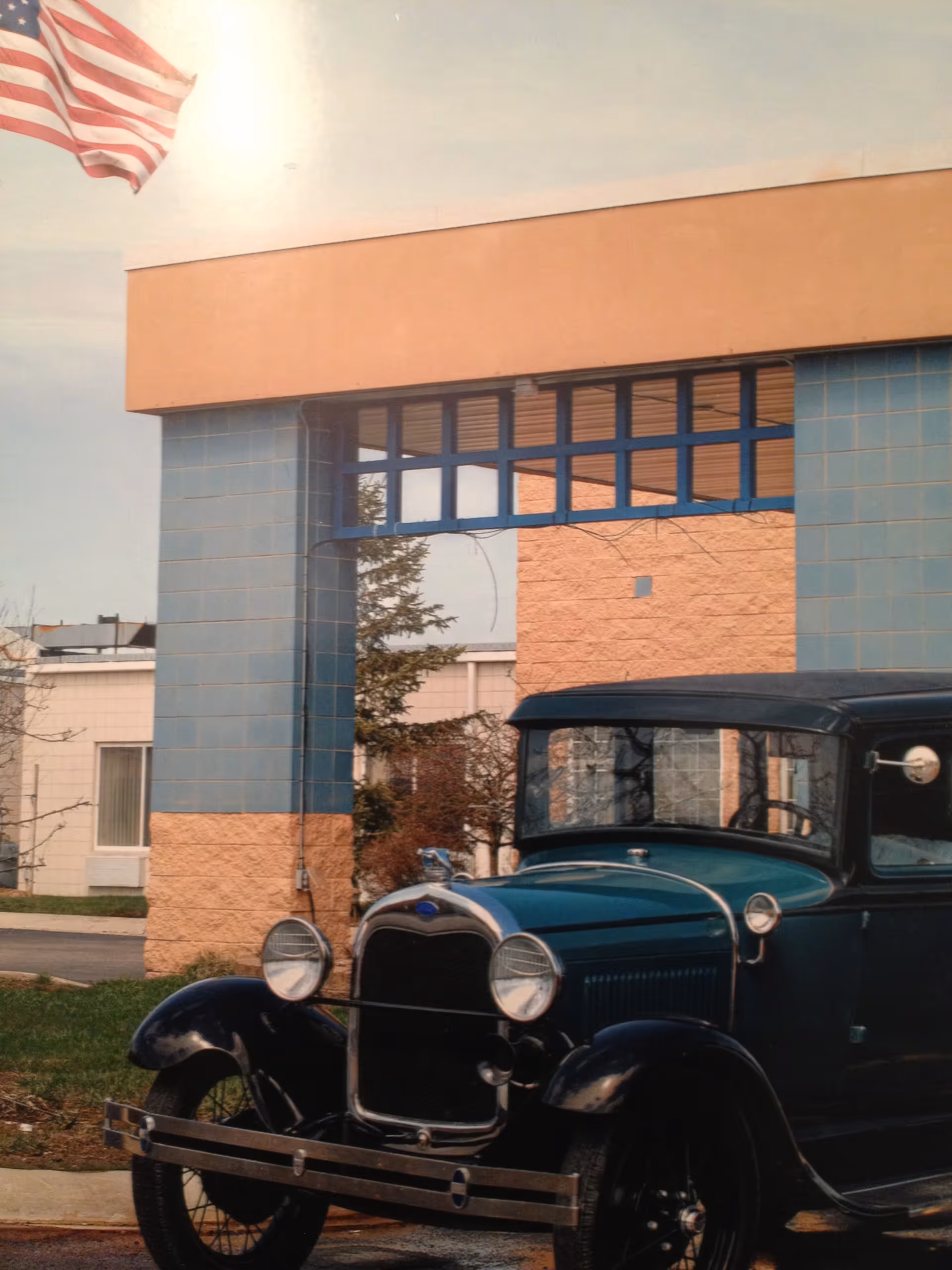 A vintage dark green and black car parked in front of a building with blue and beige tiled pillars and an American flag waving in the background.