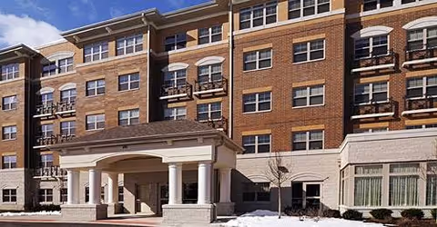 Exterior of a multi-story brick senior living building with a covered porte-cochere, balconies, and a light dusting of snow.