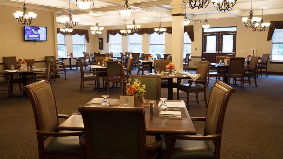 A spacious dining room with multiple wooden tables and chairs arranged neatly. Each table is set with glasses, napkins, and small floral centerpieces. Large windows with brown valances allow natural light to fill the room. Several chandeliers hang from the ceiling, providing warm lighting. A wall-mounted TV is visible on the left side of the room.