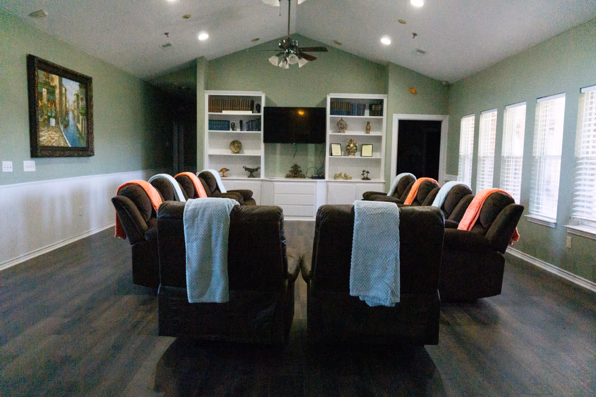 A common room with recliner chairs arranged in rows facing a wall-mounted TV and built-in shelves.