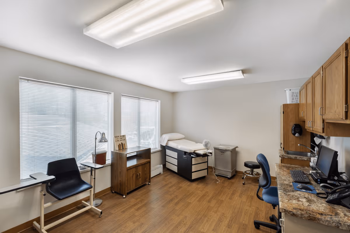 A medical examination room with two large windows covered by blinds, a black examination table with a pillow, a small fan, a trash bin, a black chair with attached arm rests, a wooden cabinet, and a countertop with a computer and office chair. The room has wooden flooring and bright overhead fluorescent lighting.