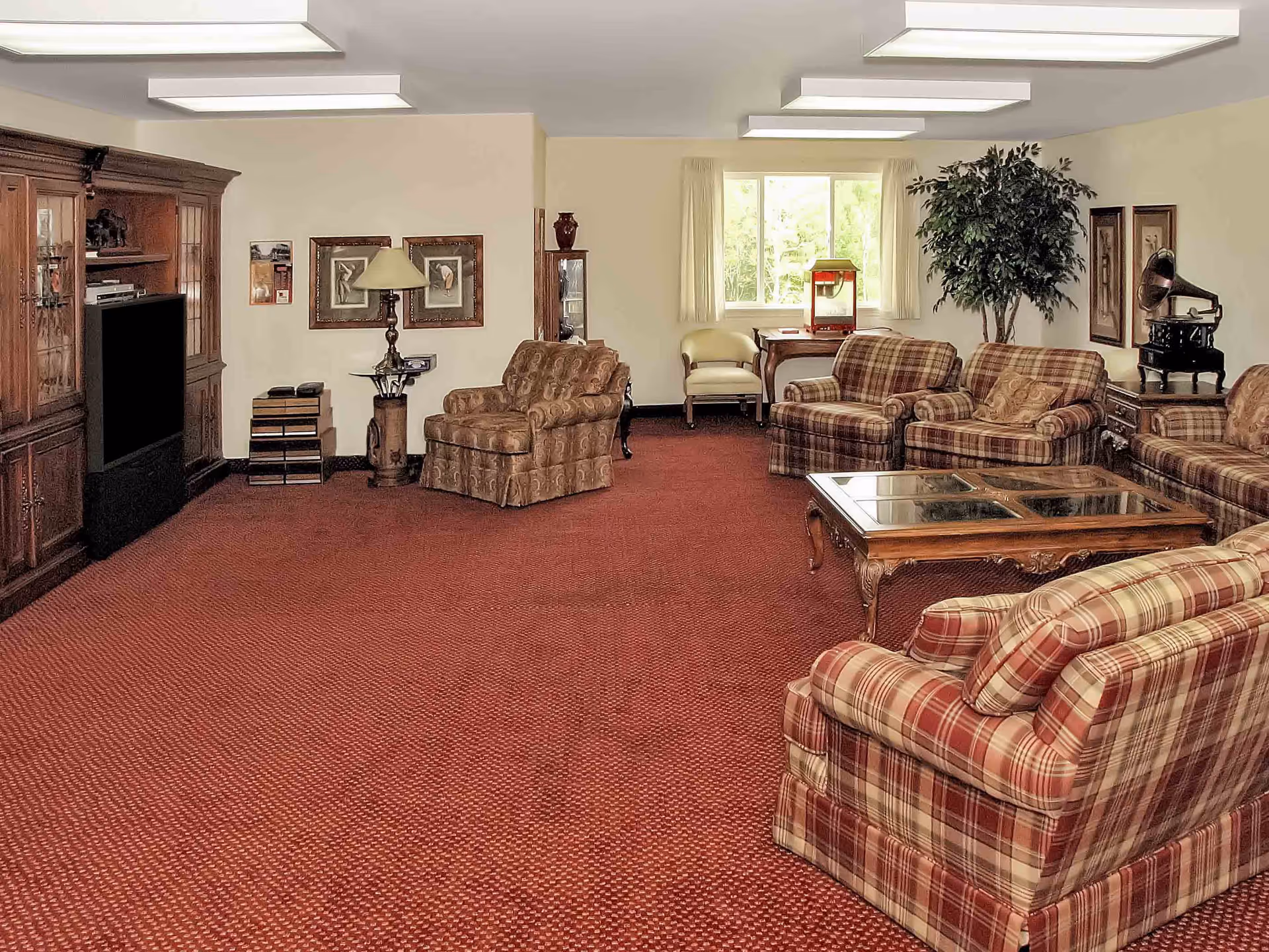 Spacious furnished lounge with plaid sofas and armchairs, a glass-top coffee table, large TV cabinet, and red carpet.