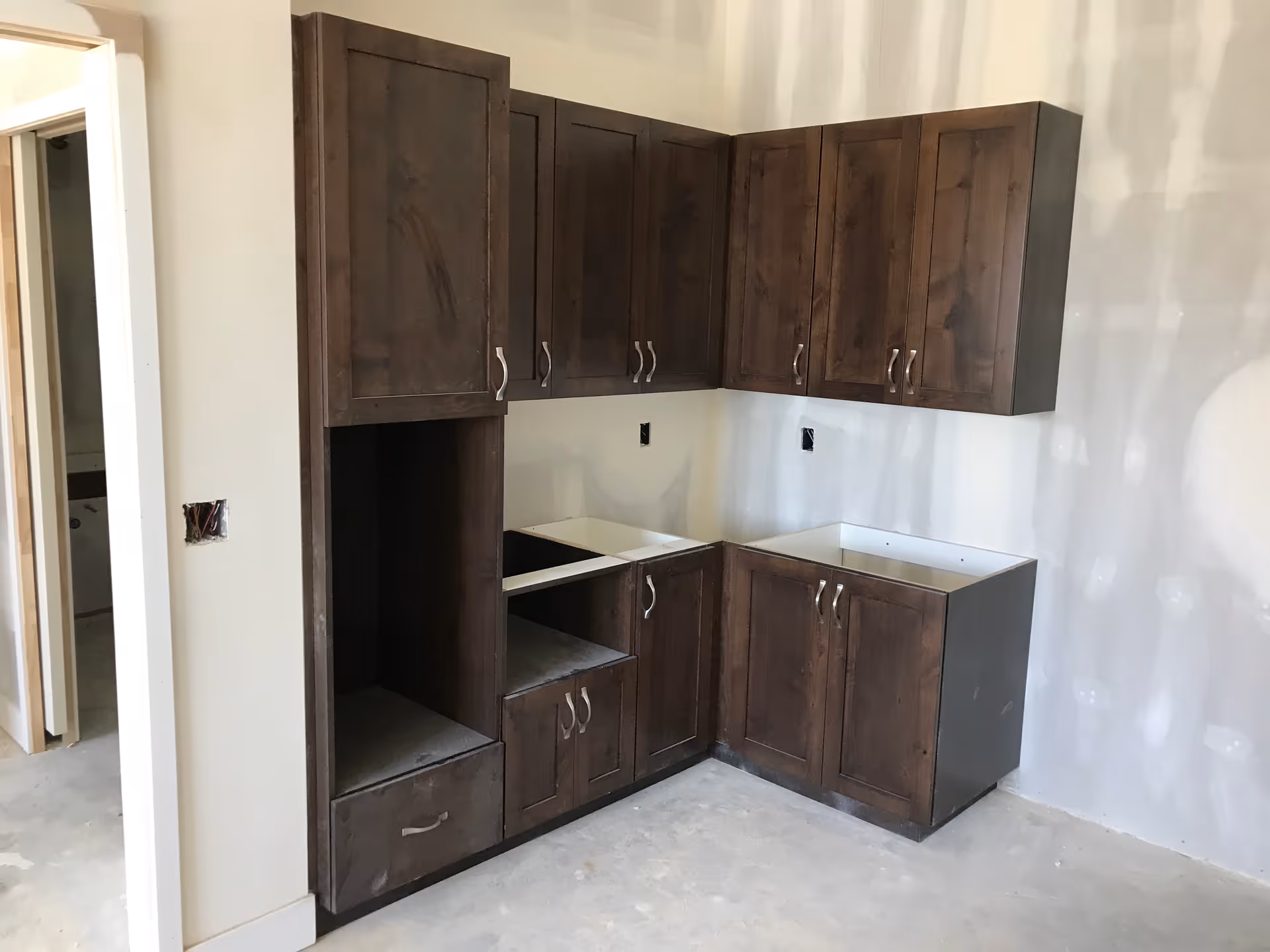 An unfinished kitchen area with dark wooden cabinets installed on the walls and floor. The room has unpainted drywall and no appliances or countertops yet. There is an open doorway leading to another room.
