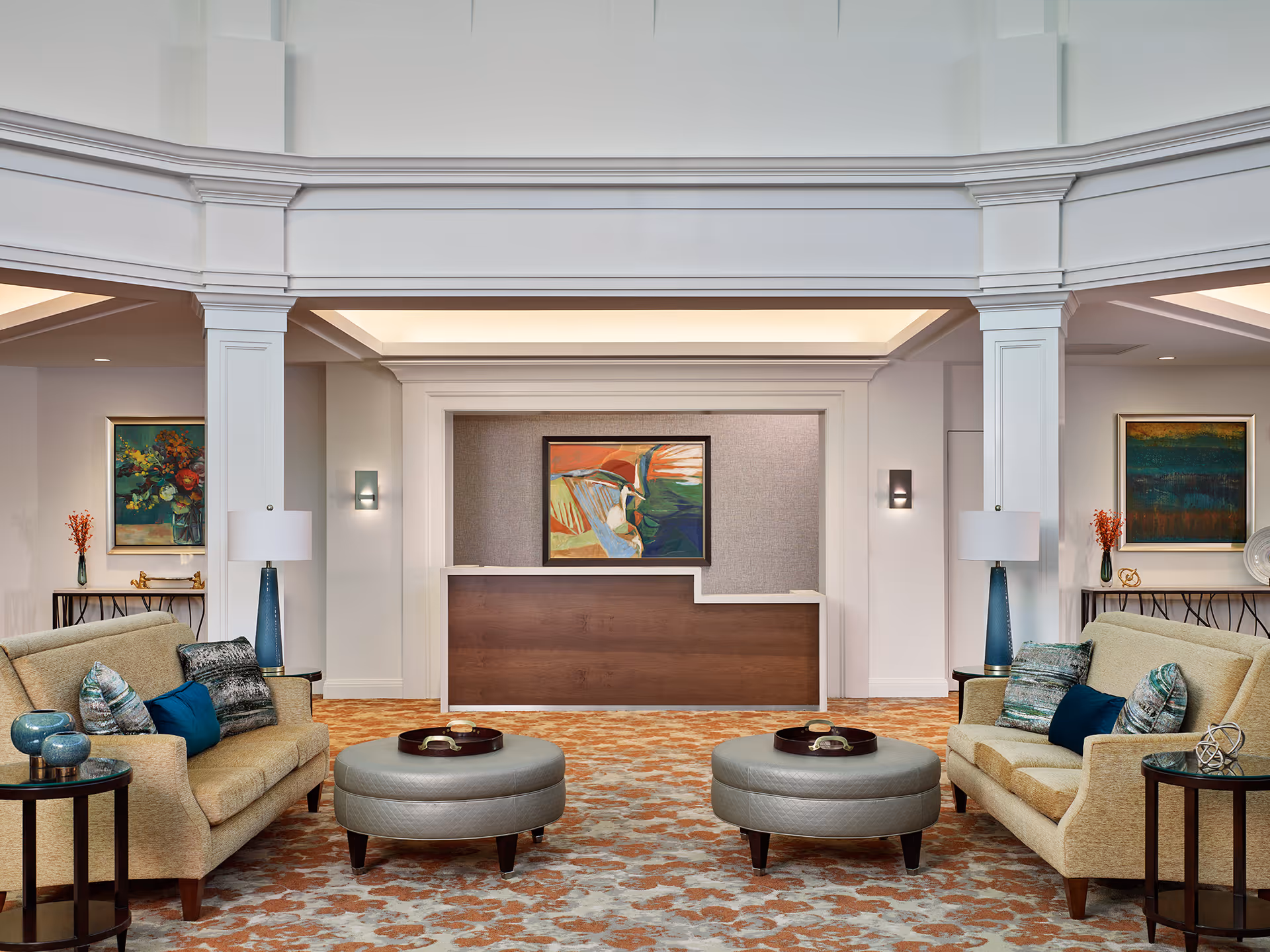 A spacious and elegant lobby area with two beige sofas facing each other, each adorned with decorative pillows. Between the sofas are two round gray ottomans with trays on top. The room features a patterned carpet in shades of orange and beige, two side tables with blue lamps, and colorful abstract paintings on the walls. The reception desk is centered in the background beneath a high ceiling with white architectural details.