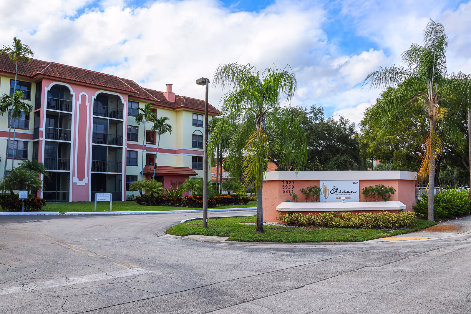 Exterior view of Elison Independent Living of Lake Worth facility showing a multi-story residential building with balconies, palm trees, and a sign with the facility name surrounded by landscaping under a partly cloudy sky.