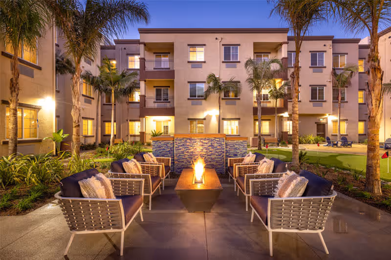 Outdoor courtyard area of a senior living facility at dusk with modern patio furniture arranged around a rectangular fire pit. The courtyard is surrounded by a three-story building with lit windows and palm trees, creating a warm and inviting atmosphere.