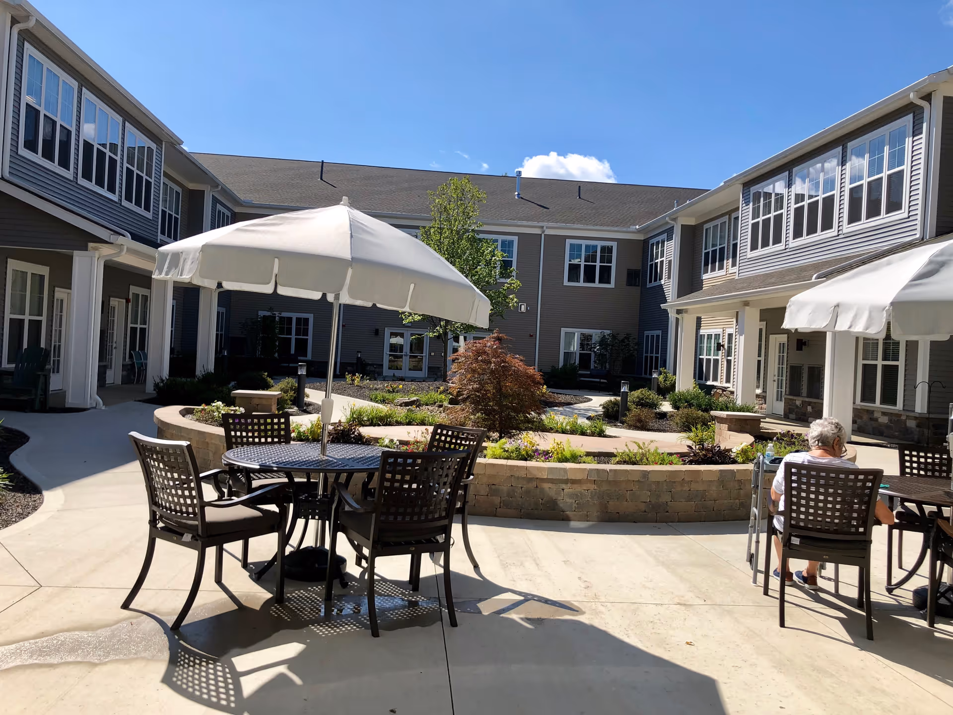 Outdoor courtyard area at Vitalia Senior Residences Westlake with patio tables and chairs under large white umbrellas, surrounded by a two-story building with many windows. There is a small garden with plants and a tree in the center, and an elderly person sitting at one of the tables.