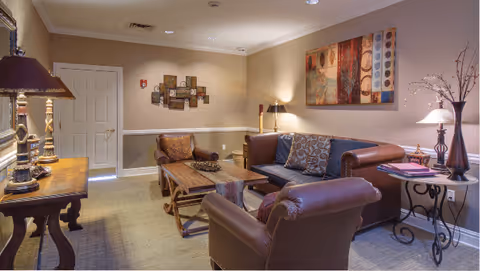 Communal sitting area with leather sofa and armchairs around a wooden coffee table, lamps, and wall art.
