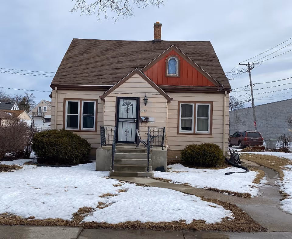 Front view of a small beige house with a brown roof, central front steps and door, and a snow-covered lawn.