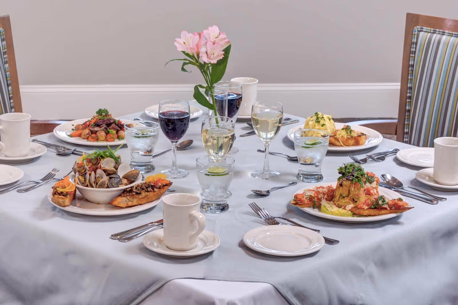 A round dining table set with plated seafood dishes, wine glasses, water glasses, coffee cups, and a vase of pink flowers.