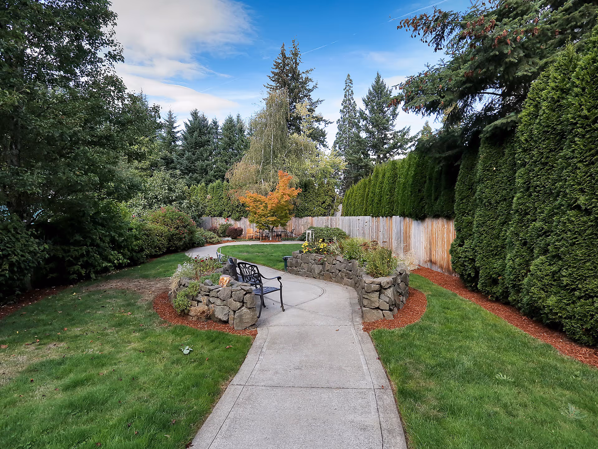 A winding concrete pathway in a garden area surrounded by green grass, tall evergreen trees, and various shrubs. There is a black metal bench placed along the path next to a low stone wall with plants. The sky is partly cloudy with blue patches visible.