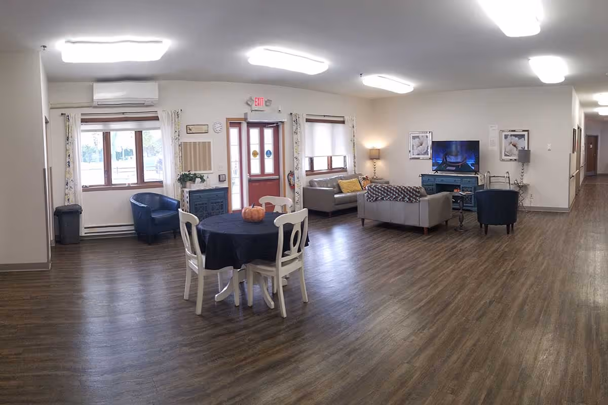 A spacious common area in a senior living facility with wooden flooring, a round table covered with a dark cloth and four white chairs around it. There are two gray sofas, a blue armchair, a TV on a blue cabinet, and lamps providing warm lighting. Windows with curtains and a red exit door are visible in the background.