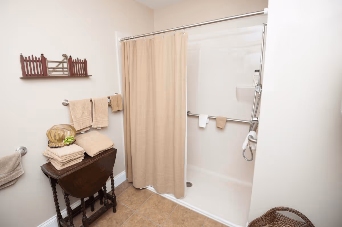 A bathroom with a walk-in shower featuring a beige shower curtain and grab bars. There are beige towels hanging on towel racks and folded on a small wooden table. The floor is tiled, and a decorative wooden wall shelf is mounted on the wall.