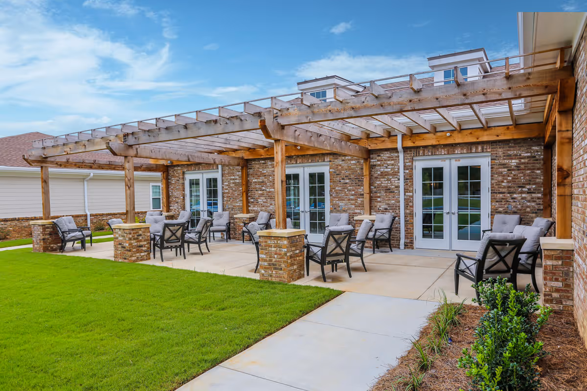 Covered outdoor patio with cushioned chairs and tables under a wooden pergola beside a brick building.