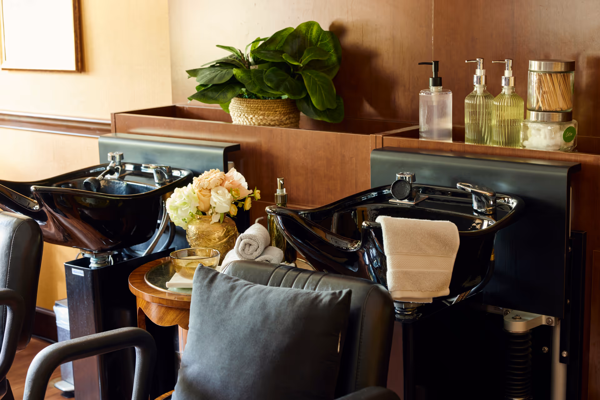 Interior view of a hair washing station with two black shampoo bowls and chairs. A small wooden table between the stations holds a vase with flowers, rolled towels, and a glass bowl. On the counter behind the bowls are bottles of soap or shampoo, cotton balls, and toothpicks. A green potted plant is visible in the background.