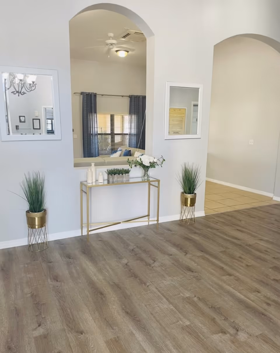 Interior view of a senior living facility with light gray walls and wood flooring. A gold-framed console table holds decorative items including white bottles, a small green plant, and a white vase with flowers. Two gold plant stands with green plants flank the table. There are two arched openings in the wall, one showing a glimpse of a living room with a ceiling fan, window with blue curtains, and a sofa.