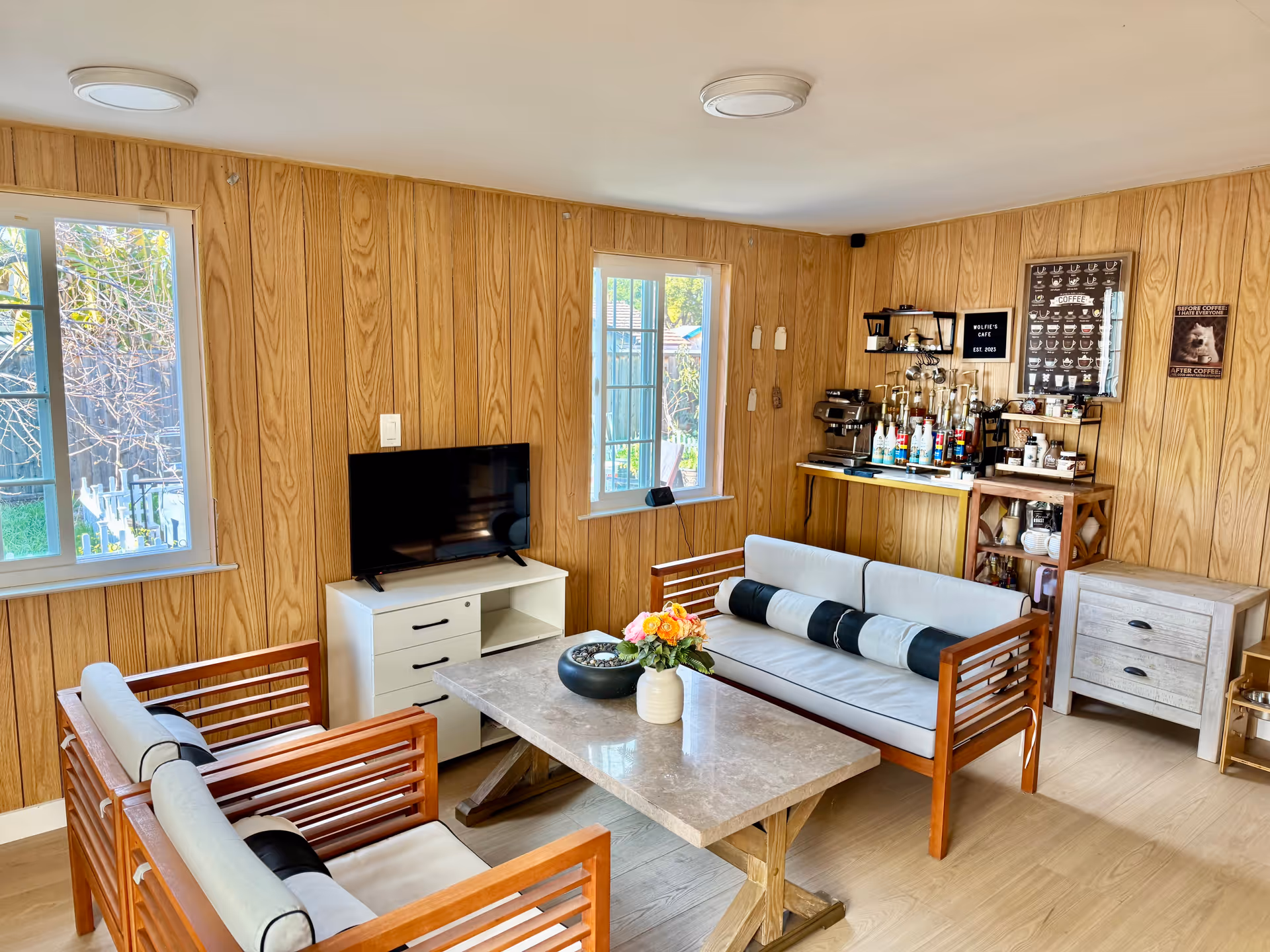 A cozy living room with wood-paneled walls, two wooden sofas with light cushions and black cylindrical pillows, a marble coffee table with a flower vase and decorative bowl, a small white TV stand with a flat-screen TV, and a coffee station with various syrups and coffee equipment on shelves. Two windows let in natural light, and there is a small wooden cabinet on the right side.