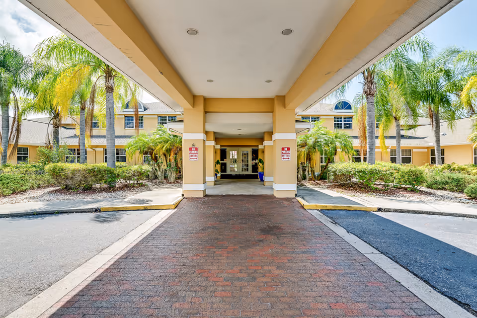Wide entrance to a yellow senior living building with a porte-cochere and palm trees.
