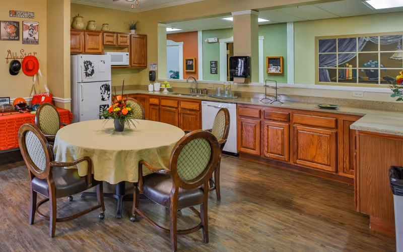 A kitchen and dining area with wooden cabinets, a white refrigerator, dishwasher, and a round table covered with a beige tablecloth surrounded by four wooden chairs with green cushions. The room has wood flooring, a countertop with a sink, and decorative items including a flower vase on the table and wall art.