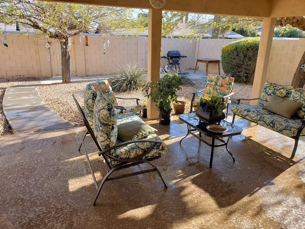 Covered outdoor patio area with floral cushioned chairs and a glass-top coffee table with potted plants. The patio overlooks a garden with gravel ground, a tree with hanging decorations, a barbecue grill, and a small table against a beige brick wall fence.