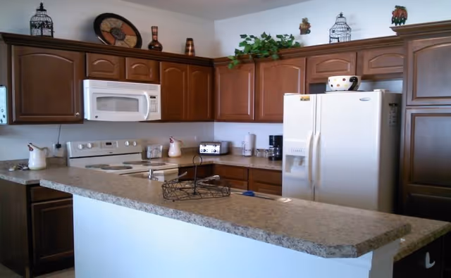 A kitchen with brown wooden cabinets, a white microwave above a white stove, a white refrigerator, and a countertop with a sink and a wire basket. Decorative items are placed on top of the cabinets, including a plant and ceramic pieces.