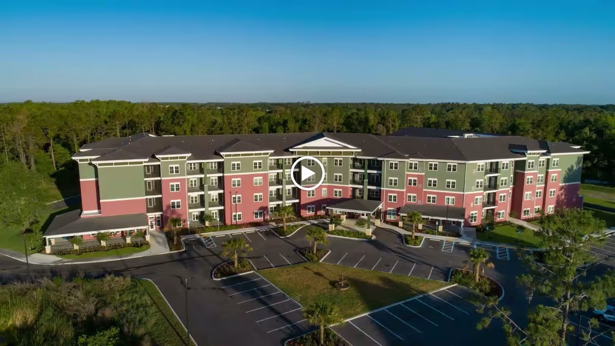 Aerial view of a large, multi-story senior living facility building with green and red exterior walls, surrounded by parking lots and greenery under a clear blue sky.