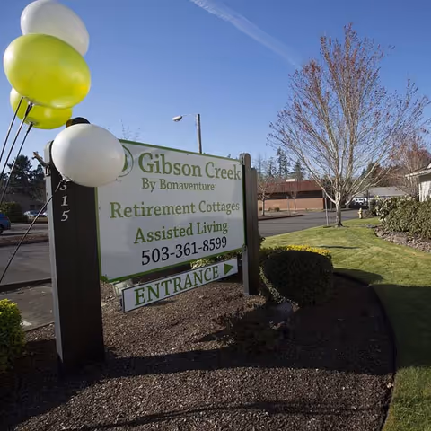 Outdoor view of a sign for Gibson Creek by Bonaventure, advertising Retirement Cottages and Assisted Living with a phone number and an entrance arrow. The sign is decorated with green and white balloons and is situated on a landscaped area with grass, bushes, and trees under a clear blue sky.