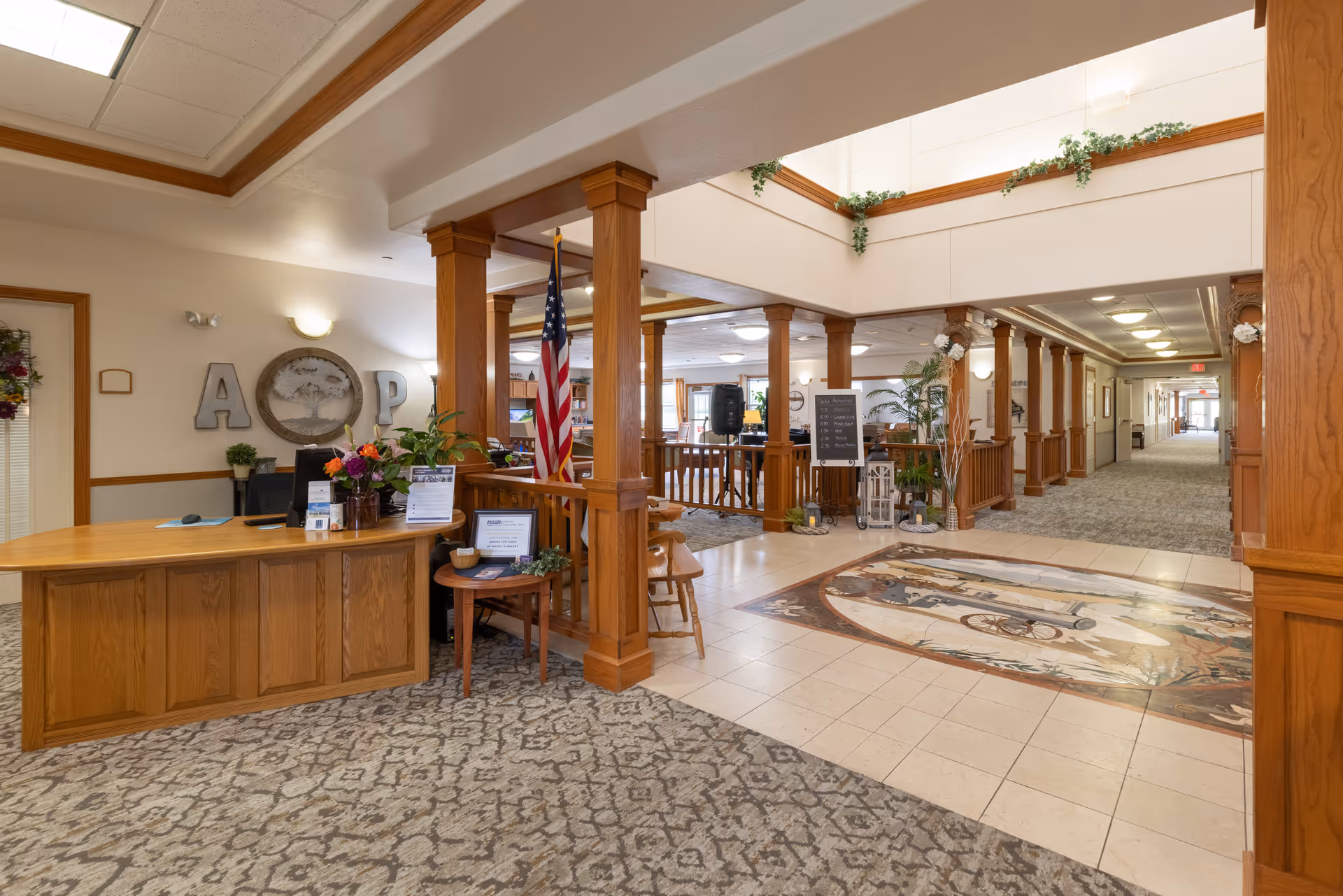 Spacious senior living facility lobby with a wooden reception desk, columns, American flag, seating area and decorative tiled floor.