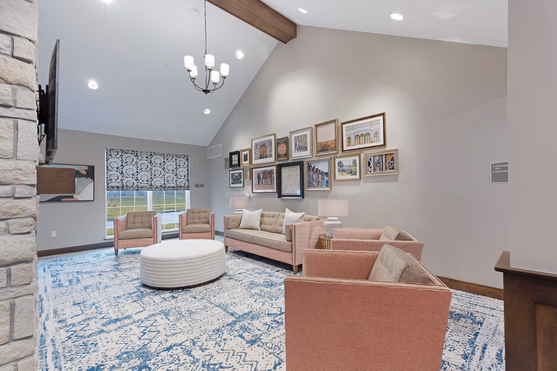 A bright and spacious senior living facility lounge area with a high vaulted ceiling, a chandelier, and a large window with patterned blinds. The room features a beige sofa, two matching armchairs, a round white ottoman, and a blue and white patterned carpet. The walls are decorated with a collection of framed artwork.