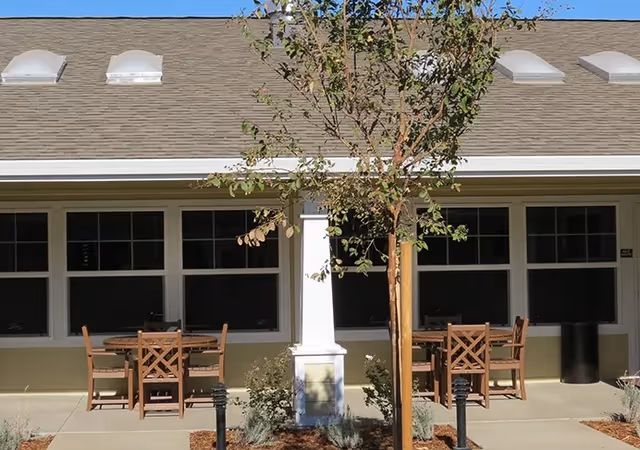 Outdoor patio with wooden tables and chairs in front of a single-story building with multiple windows and a small tree.