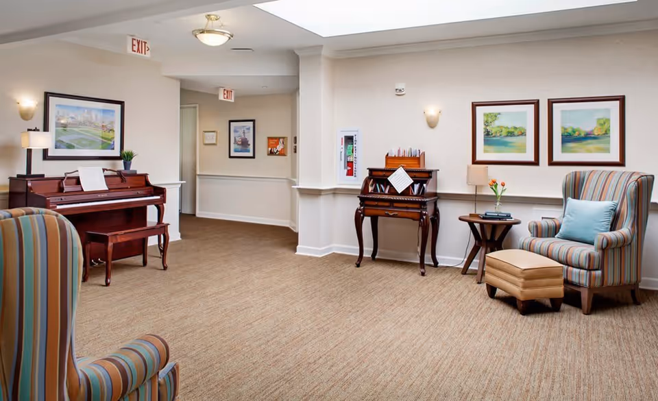 A cozy interior common area featuring a wooden piano with sheet music, a small antique-style desk with papers, a side table with a lamp and flowers, and two striped armchairs with a matching ottoman. The walls are decorated with framed landscape paintings, and the carpet is beige. There are exit signs visible in the hallway.