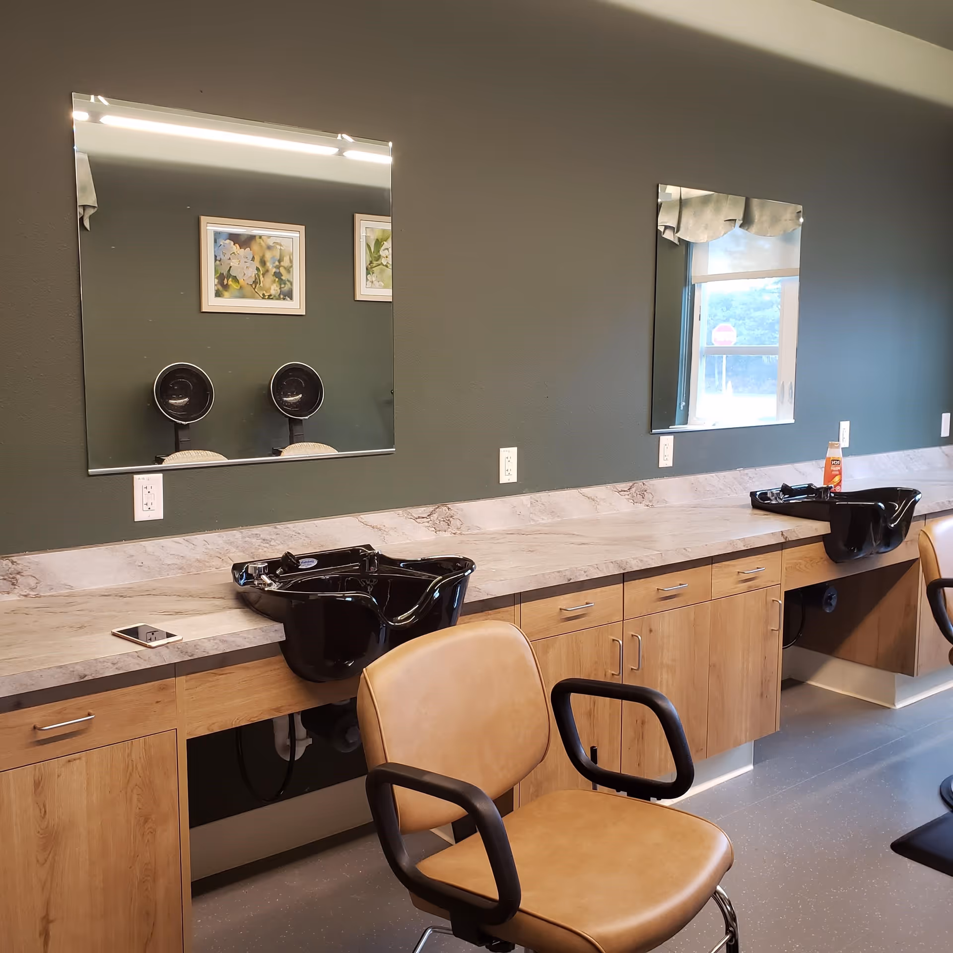 Interior of a salon area with two black hair washing sinks, two mirrors mounted on a dark green wall, wooden cabinets below the countertop, and two tan salon chairs with black armrests. There are framed pictures reflected in one mirror and a window reflected in the other.