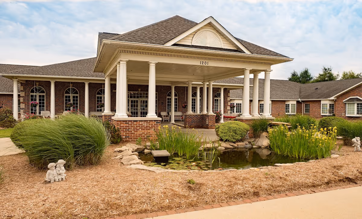 Front exterior view of a senior living facility with a covered entrance supported by white columns, brick walls, large windows, landscaped garden with tall grasses, yellow flowers, a small pond with lily pads, and decorative statues.