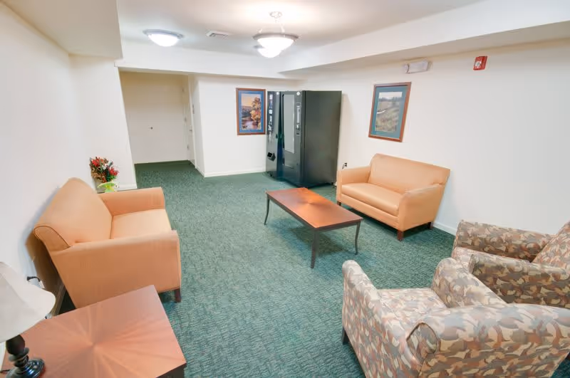 A small lounge area with two beige sofas, two patterned armchairs, a wooden coffee table, and a vending machine against the far wall. The room has green carpet, white walls, two framed pictures, and ceiling lights.