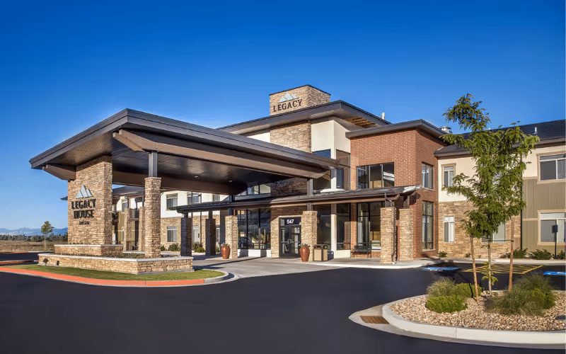 Exterior view of Legacy House of Park Lane, a modern senior living facility with stone and brick facade, large covered entrance, and clear blue sky.