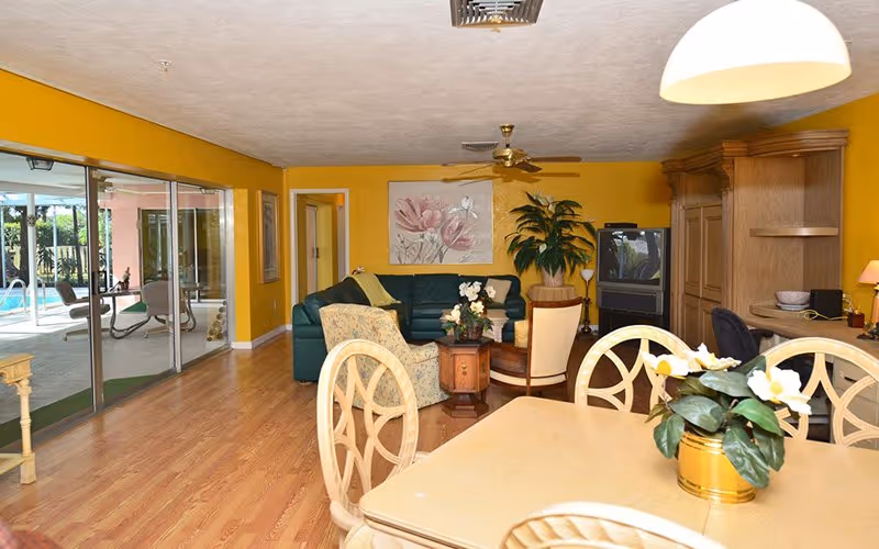 Open living and dining room with yellow walls, a dining table in the foreground, a green sofa and TV in the seating area, and sliding glass doors opening to a pool.