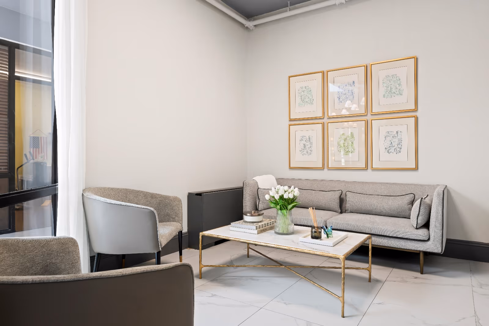 A modern living room with a light gray sofa, two gray armchairs, and a rectangular coffee table with a gold frame and white marble top. The coffee table holds a vase of white tulips, books, and decorative items. Six framed abstract artworks hang on the light gray wall above the sofa. The floor is white tile, and a large window with sheer white curtains is on the left side.