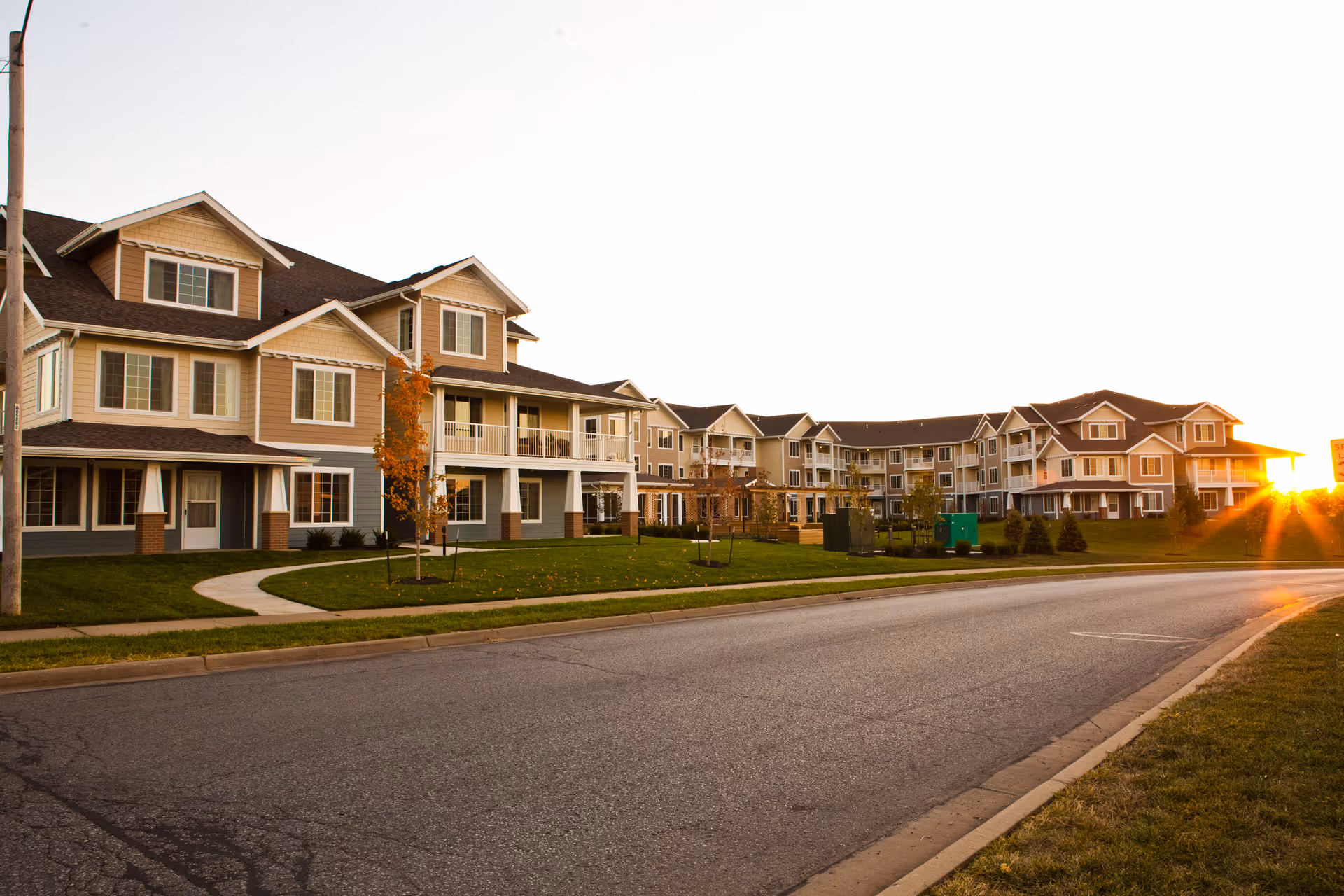 Exterior view of a large, multi-story residential building complex with beige siding and white trim, situated along a paved road with a grassy lawn and young trees, captured at sunset with the sun low on the horizon.