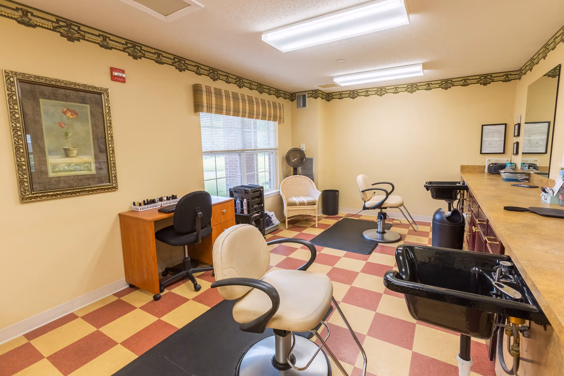 Interior of a salon room with two beige salon chairs, two black hair washing sinks, a wooden desk with a black office chair, a wicker chair near a window with plaid valance, and framed artwork on the walls. The floor has a red and yellow checkered pattern.