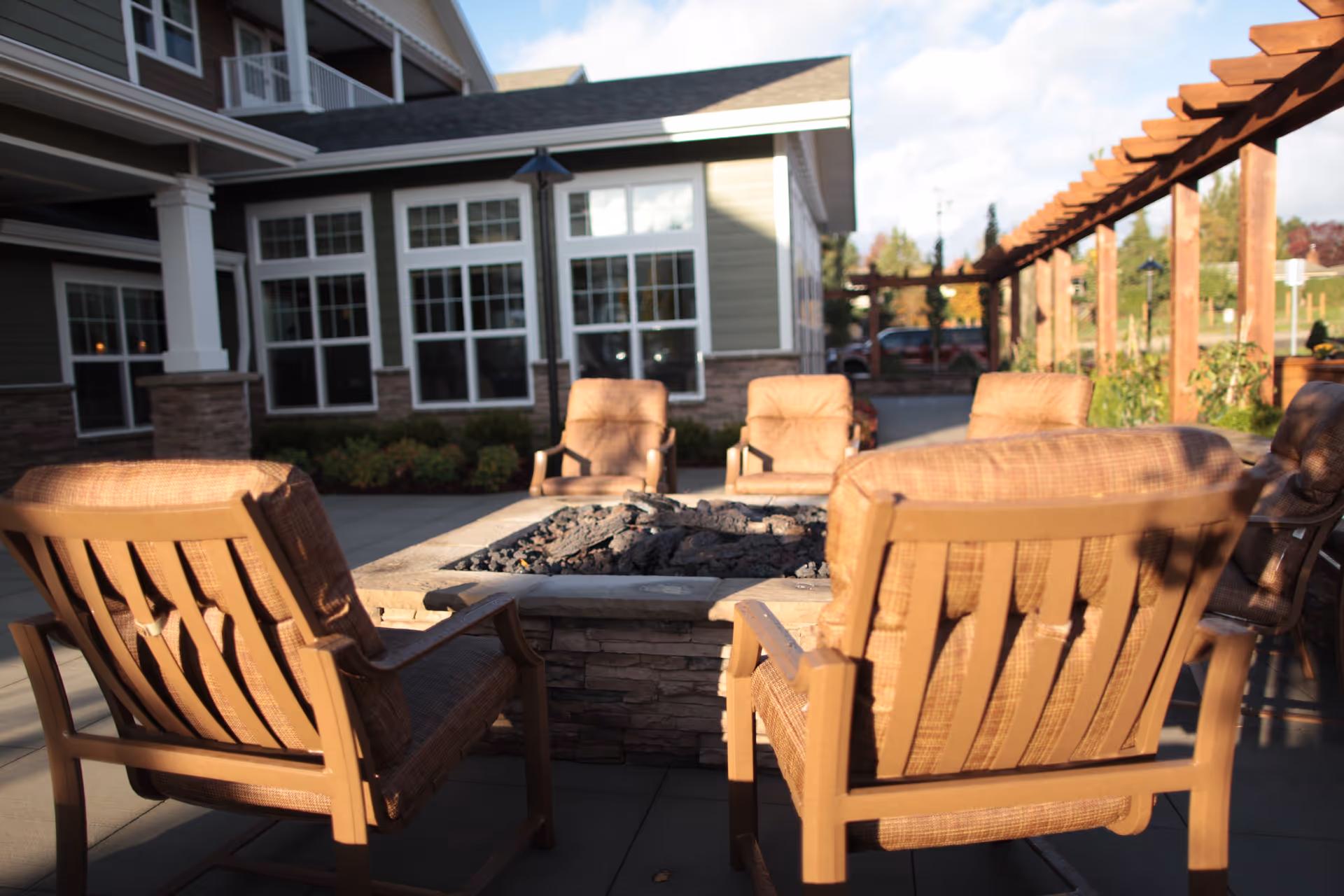 Cushioned patio chairs arranged around a square fire pit on an outdoor patio in front of a retirement home building.