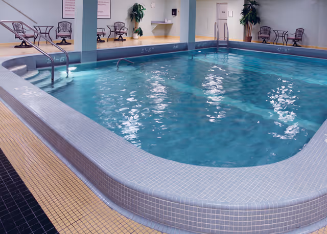 Indoor swimming pool with clear blue water surrounded by light blue tiled edges. There are metal handrails for entering the pool and several chairs and tables along the walls. Potted plants and wall-mounted items are visible in the background.